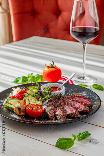 chopped medium rare steak with grilled vegetables. The dish is served with tomato sauce and a glass of red wine. Dish on a white plate in natural light.