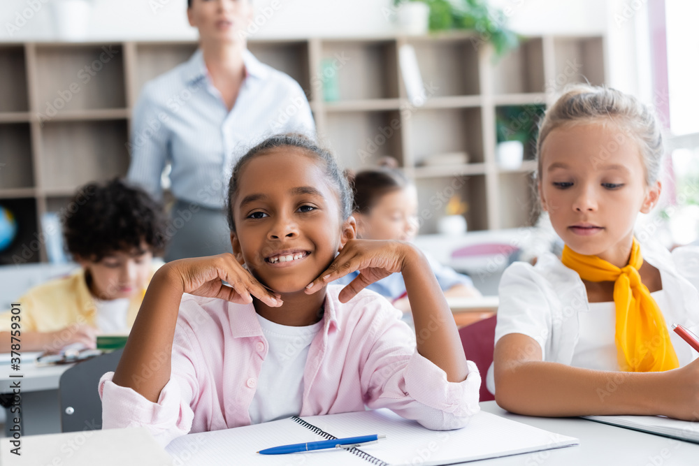 Selective focus of african american schoolgirl looking at camera near classmates and teacher in school Selective focus of african american schoolgirl looking at camera near classmates and teacher in school