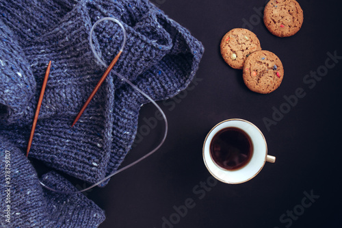 Blue knit sweater, knitting needles, porcelain gold cup with coffee and flavored cookies with colored drops on black background. Knitwear as a concept of female hobby