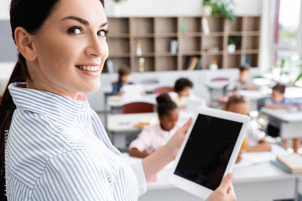 selective focus of teacher holding digital tablet with blank screen and looking at camera near multicultural pupils in classroom selective focus of teacher holding digital tablet with blank screen and looking at camera near multicultural pupils in classroom
