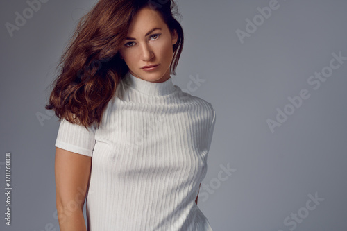 Sexy attractive young woman with shoulder length auburn hair wearing a high necked white summer top giving the camera a sultry sensual look over a grey background with copy space photo