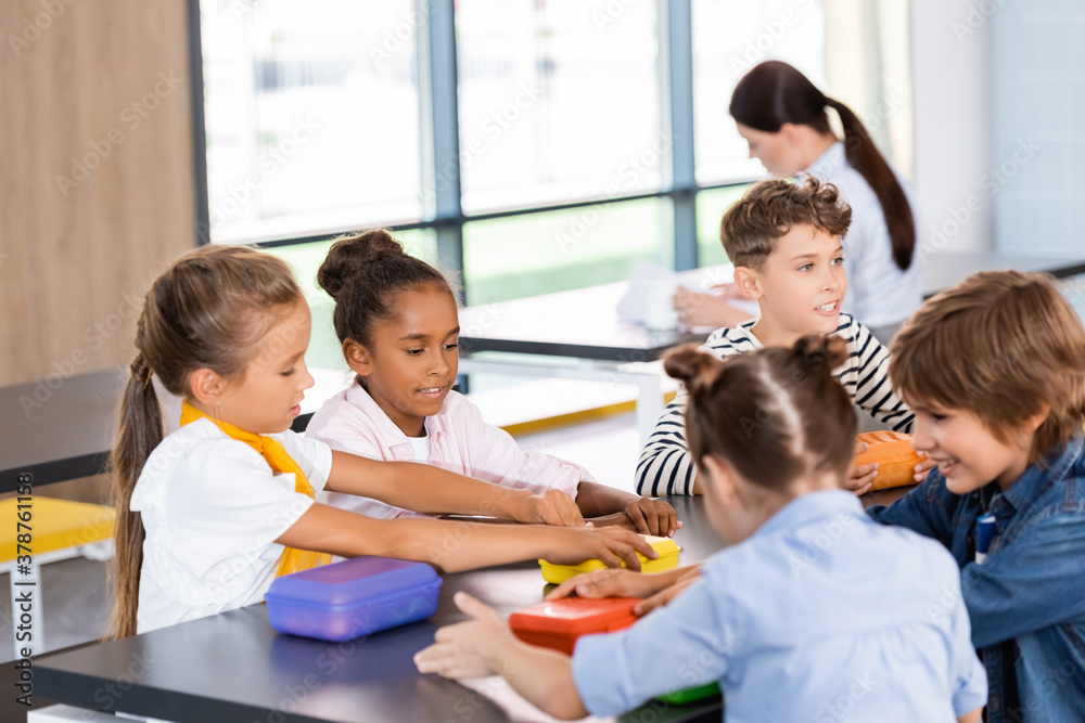 selective focus of multiethnic classmates sitting in school canteen near lunch boxes selective focus of multiethnic classmates sitting in school canteen near lunch boxes