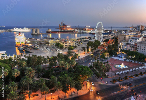 Malaga, Spain: seafront and harbour at sunset with ferris wheel at the harbour entrance. In the background the harbour in full operation with a freighter and harbour cranes.