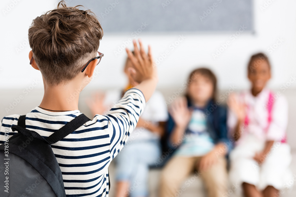 Obraz premium Selective focus of schoolboy with backpack waving hand at multiethnic classmates