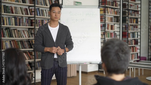 Young asian man in eyeglasses standing near white board on background of bookshelves, young male teacher giving a lecture in the library 