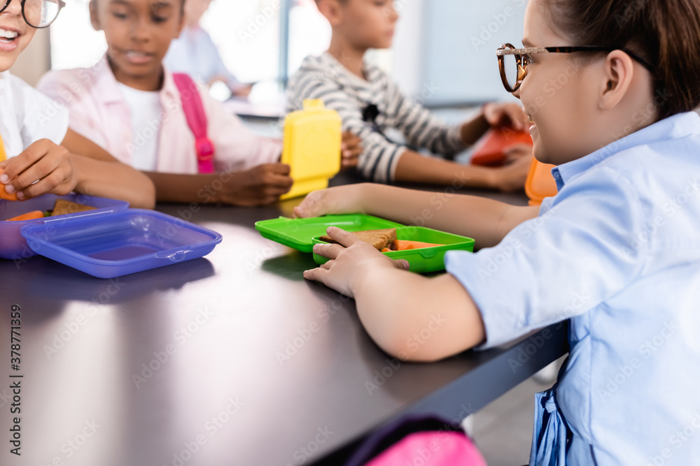 selective focus of multiethnic classmates sitting in school eatery near lunch boxes selective focus of multiethnic classmates sitting in school eatery near lunch boxes