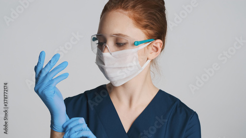 Young doctor in protective eyeglasses and mask wearing medical gloves on camera over white background