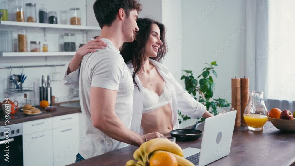 Seductive woman with pancakes in pan embracing man near laptop on table Seductive woman with pancakes in pan embracing man near laptop on table