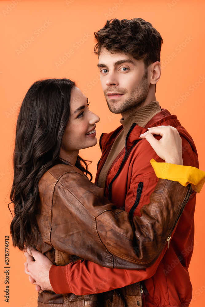 young brunette woman hugging stylish man in jacket isolated on orange young brunette woman hugging stylish man in jacket isolated on orange