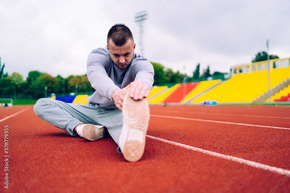 Determined caucasian male slimming and training body shape on morning workout on stadium, serious 20s man jogger stretching muscles of legs reaching body flexibility for good fitness results
