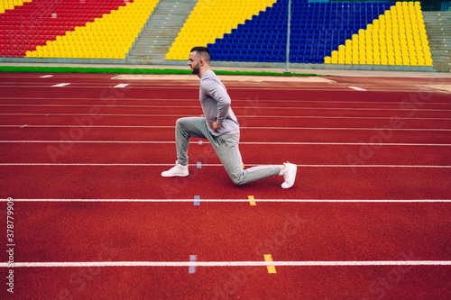 Serious caucasian male runner squatting training lower body muscles enjoying morning training on stadium, determined hipster guy having workout for slimming and losing weight for flexible body