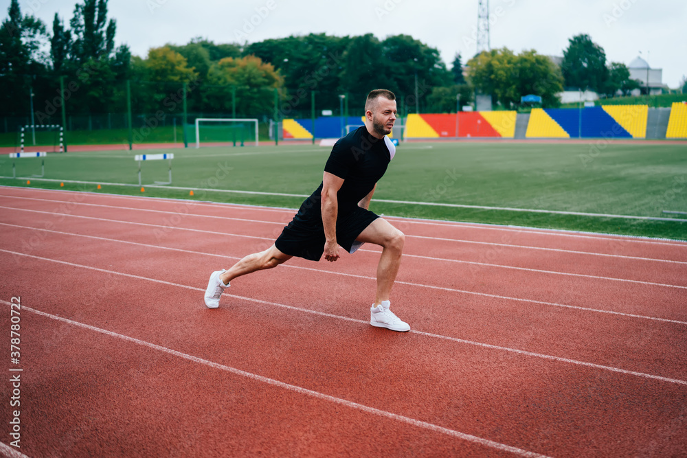 Determined caucasian man in black tracksuit doing squats for training engs warming body on training, male athlete in good body shape training flexibility and physical strength on stadium workout