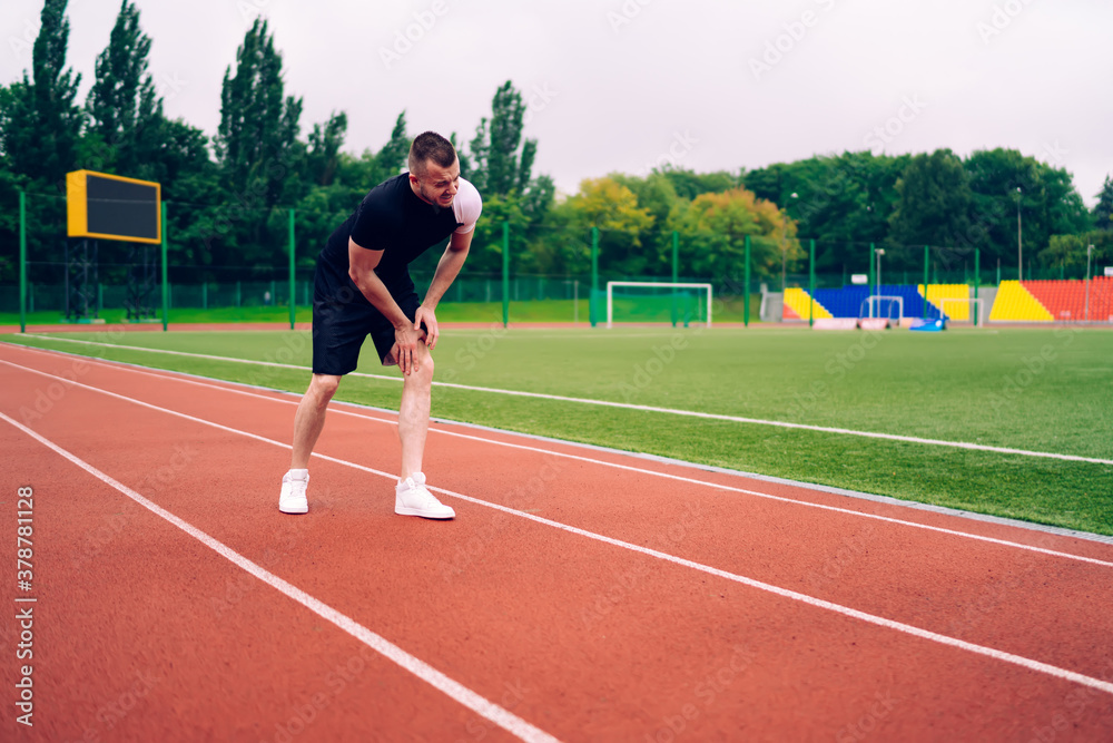 Used disappointed caucasian male runner feeling terrible pain in knee after injury need help on stadium, young man jogger having trouble with knee and knee can't walk after accident during cardio
