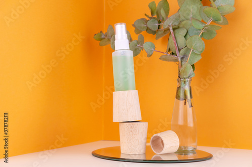 Green spray skin tonic bottle  on wooden pedestal and eucalyptus branch in glass vase on orange backdrop. Care cosmetics. Organic natural treatment for skin healing