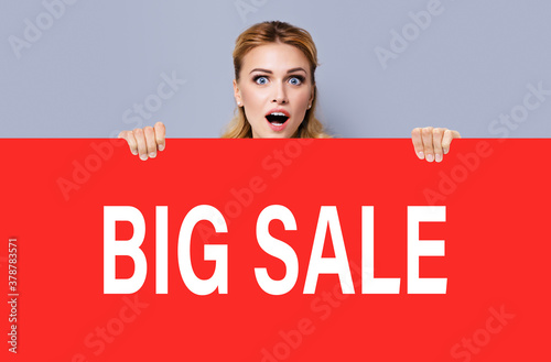 Excited surprised, shocked or very happy amazed woman with wide opened mouth and eyes, peek out from red color banner signboard with big sale sign text. Girl behind broadsheet at studio. © vgstudio