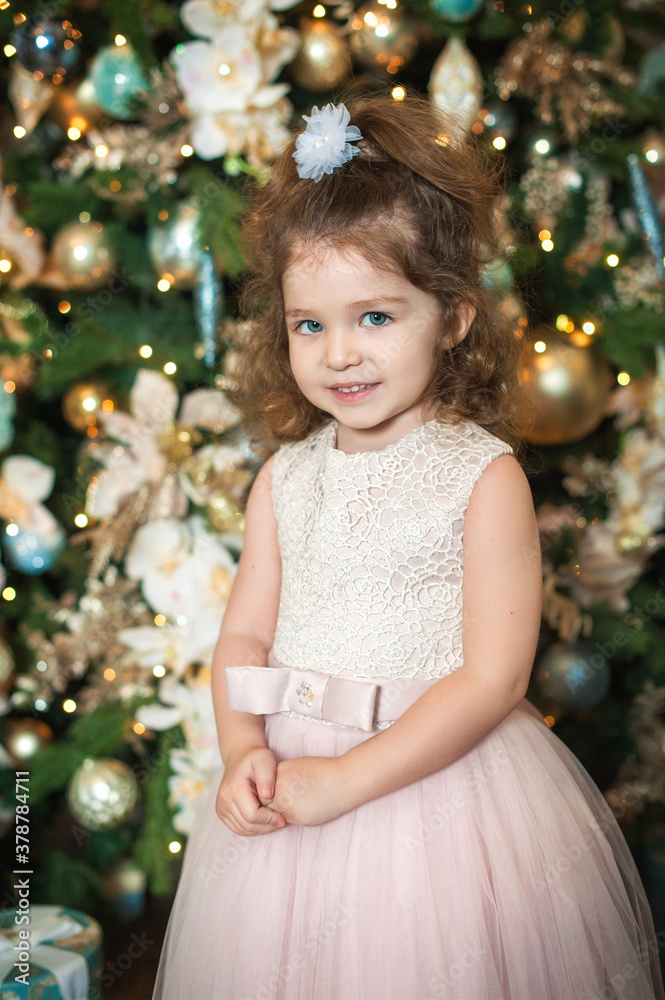 A little beautiful girl with blue eyes in a pink dress stands against the background of a decorated Christmas tree with lights and looks into the camera. Vertical photo. Children's portrait. A little beautiful girl with blue eyes in a pink dress stands against the background of a decorated Christmas tree with lights and looks into the camera. Vertical photo. Children's portrait.