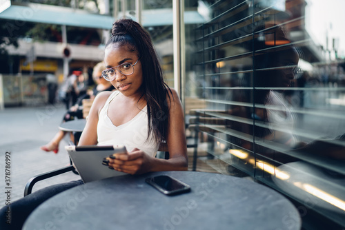Beautiful dark skinned young hipster girl in trendy eyewear looking at camera holding tablet for networking on cafe terrace, portrait of smiling african american woman typing message on touchpad
