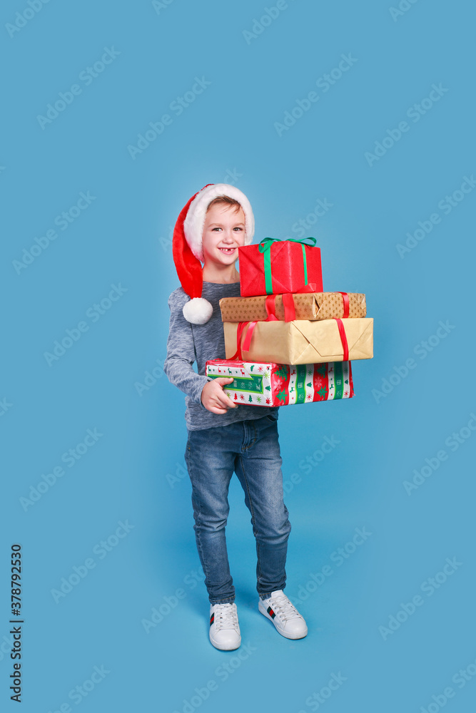 Happy smiling boy in Christmas cap holding Christmas gifts in craft paper and with a red bow on blue background. Happy smiling boy in Christmas cap holding Christmas gifts in craft paper and with a red bow on blue background.