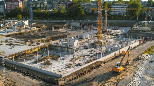 Aerial Shot of a New Constructions Development Site with High Tower Cranes Building Real Estate. Heavy Machinery and Construction Workers are Employed. City Houses in the Background.