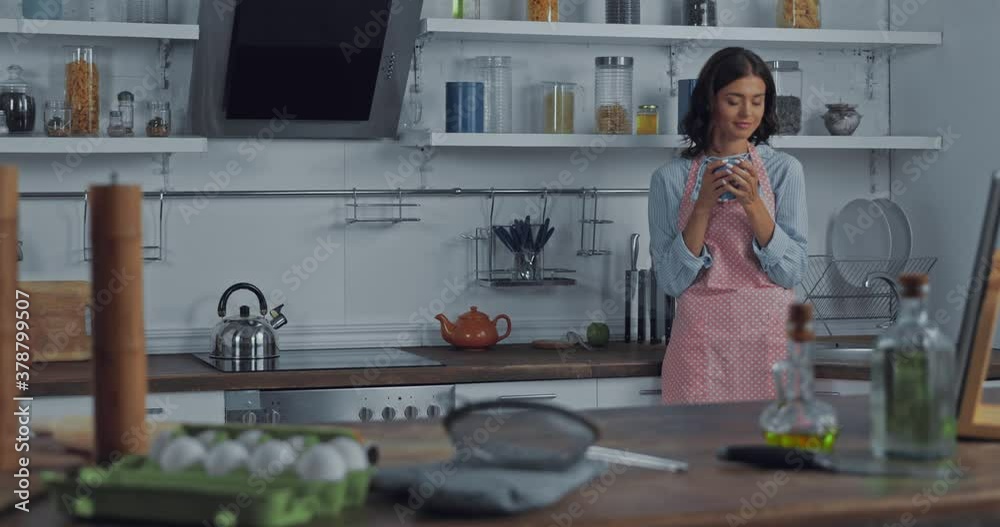 Woman in apron drinking coffee near ingredients in kitchen Woman in apron drinking coffee near ingredients in kitchen