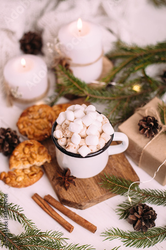 Enamelled cup of hot cocoa or coffee with marshmallows and cookies. Around the tree branches, gifts and burning candles. Christmas mood. Postcard or winter background.