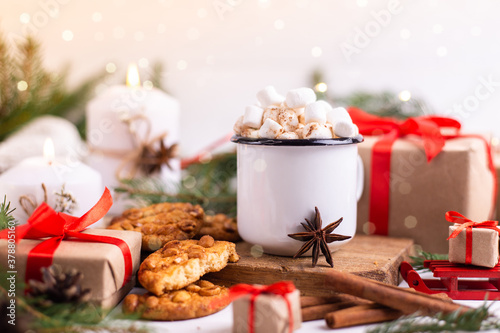 Enamelled cup of hot cocoa or coffee with marshmallows and cookies. Around the tree branches, gifts and burning candles. Christmas mood. Postcard or winter background.