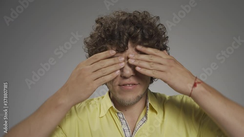 Young curly-haired guy writhes his faces on camera. He closes his eyes with his palms and lifts the corners of his lips up. High quality 4k footage. photo