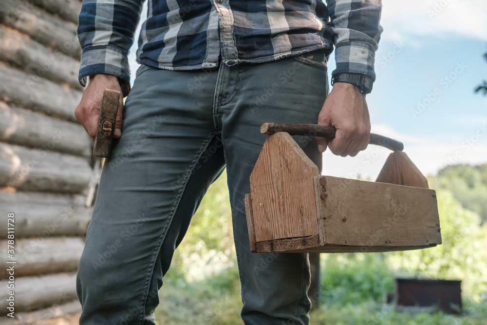 Male carpenter holds a homemade wooden box in one hand and a hammer in the other, against the backdrop of an old log barn. Male carpenter holds a homemade wooden box in one hand and a hammer in the other, against the backdrop of an old log barn.