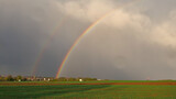 Strong double colored rainbow after thunderstorm with green fields and village in background near Plieningen, Stuttgart, Germany.