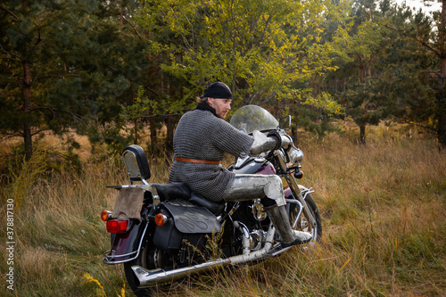 A biker in medieval knight's armor sits on a motorcycle against the background of the forest.