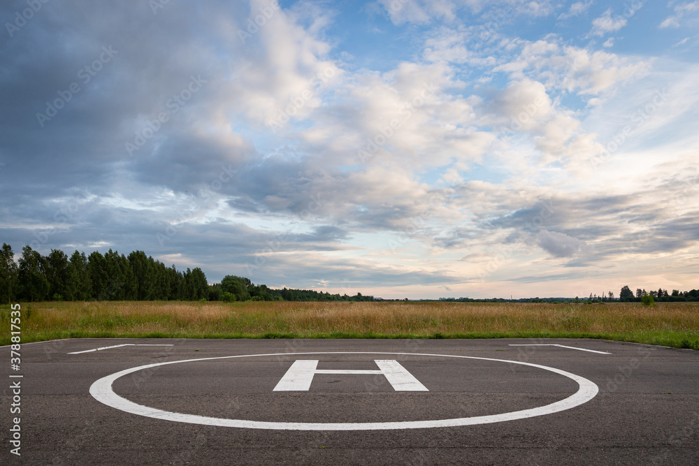 A closeup of an asphalt-covered helipad with a special symbol in the center for helicopter landing, against the backdrop of a green field and a cloudy evening sky.