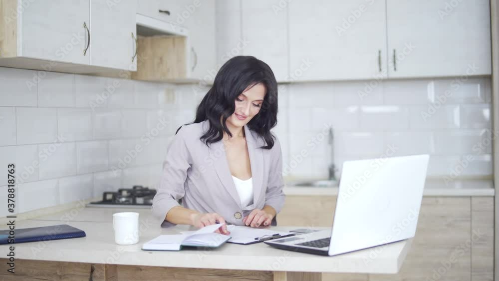 Attractive smiling business lady working with laptop and making notes at home. Happy brunette girl typing on computer and writing on paper, with coffee cup in bright white kitchen Attractive smiling business lady working with laptop and making notes at home. Happy brunette girl typing on computer and writing on paper, with coffee cup in bright white kitchen