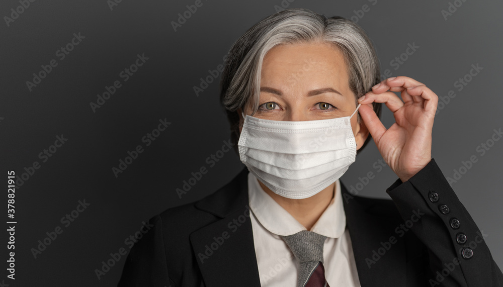 Pretty gray-haired middle-aged business woman putting on protective mask and showing ok or okey sign while posing on gray wall background. Quarantine concept. Pretty gray-haired middle-aged business woman putting on protective mask and showing ok or okey sign while posing on gray wall background. Quarantine concept.
