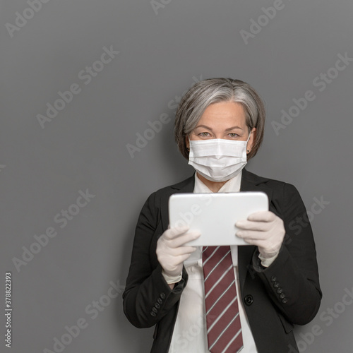 Caucasian businesswoman in protective mask works digital gadget. Gray haired lady looks at camera holding tablet before herself. High quality photo