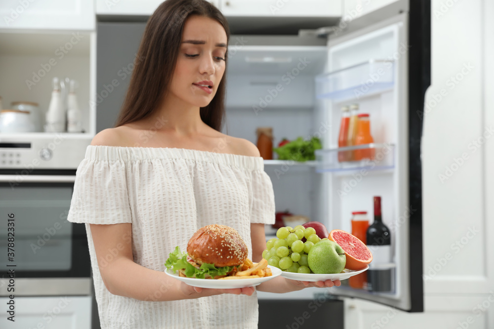 Woman choosing between fruits and burger with French fries near refrigerator in kitchen