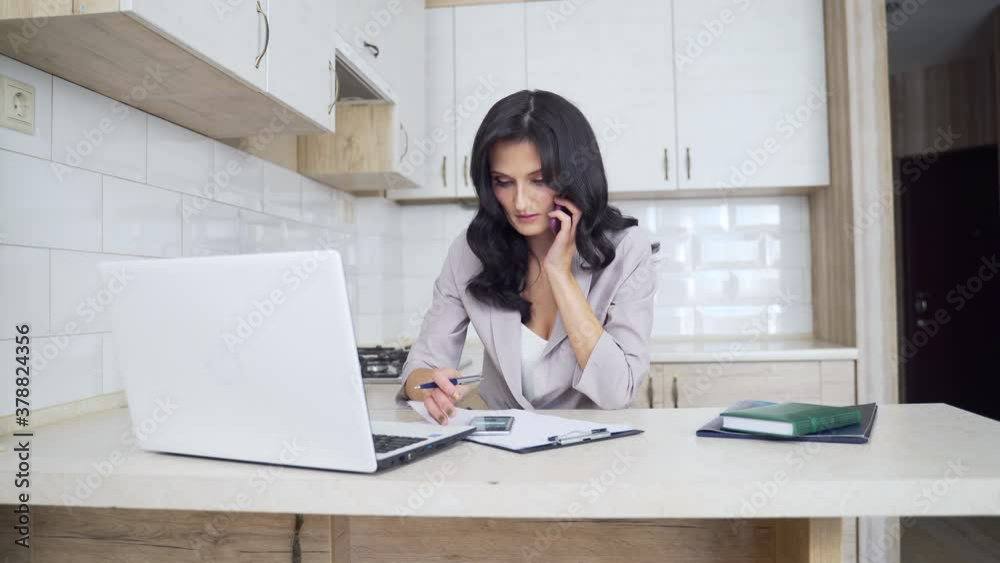 Young attractive sexy businesswoman talking on smartphone and making notes at bright kitchen. Confident brunette lady sitting and working with white laptop from home. Young attractive sexy businesswoman talking on smartphone and making notes at bright kitchen. Confident brunette lady sitting and working with white laptop from home.