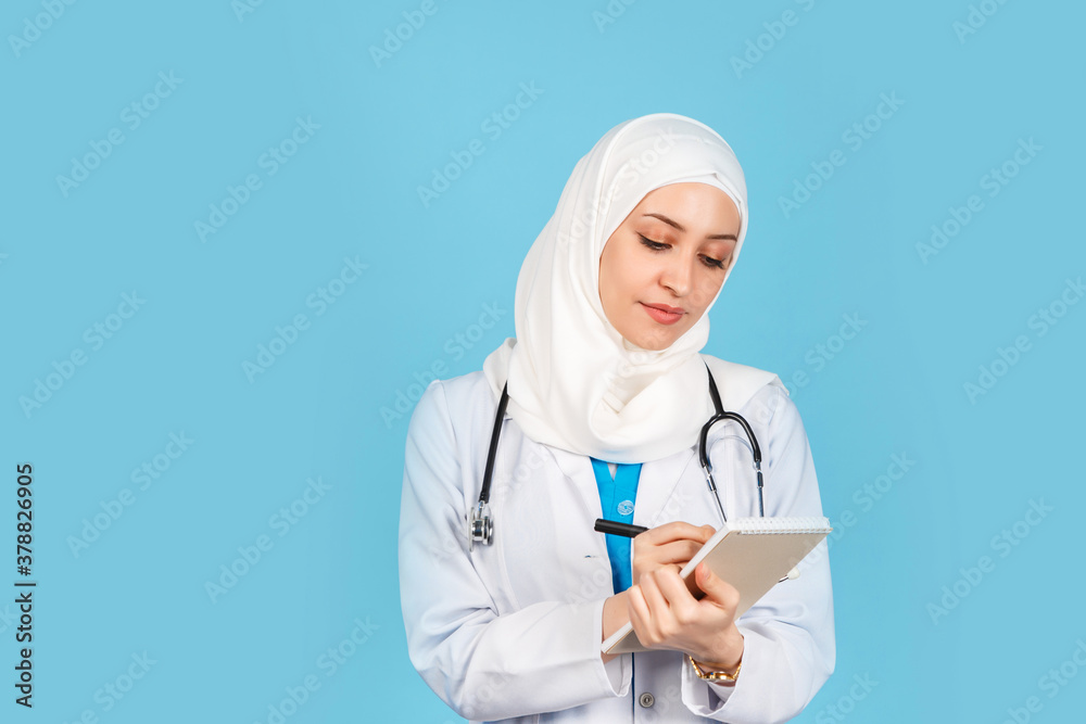 friendly, Muslim doctor or nurse woman in a hijab with a stethoscope writes a prescription on pills on a blue background. friendly, Muslim doctor or nurse woman in a hijab with a stethoscope writes a prescription on pills on a blue background.