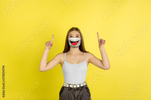 Pointing. Portrait of young girl with emotion on her protective face mask on studio background. Beautiful female model, funny expression. Human emotions, facial expression, sales, ad, healthcare