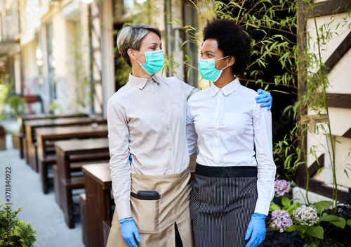 Happy waitresses wearing protective face masks and gloves while working at outdoor cafe.