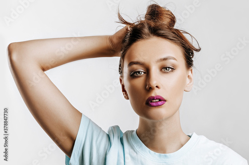 A beautiful brunette girl holds her hair and makes her own hairstyle. Young model with long hair, fashion makeup with purple lips on an gray background. The woman seriously looking at the camera.