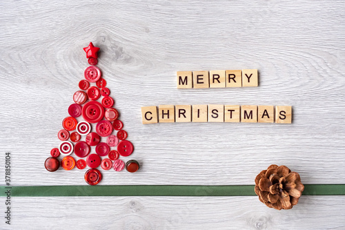 Christmas tree made of red buttons and the inscription of wooden letters merry christmas on a white wooden background. below is a green ribbon and a bump. top view.