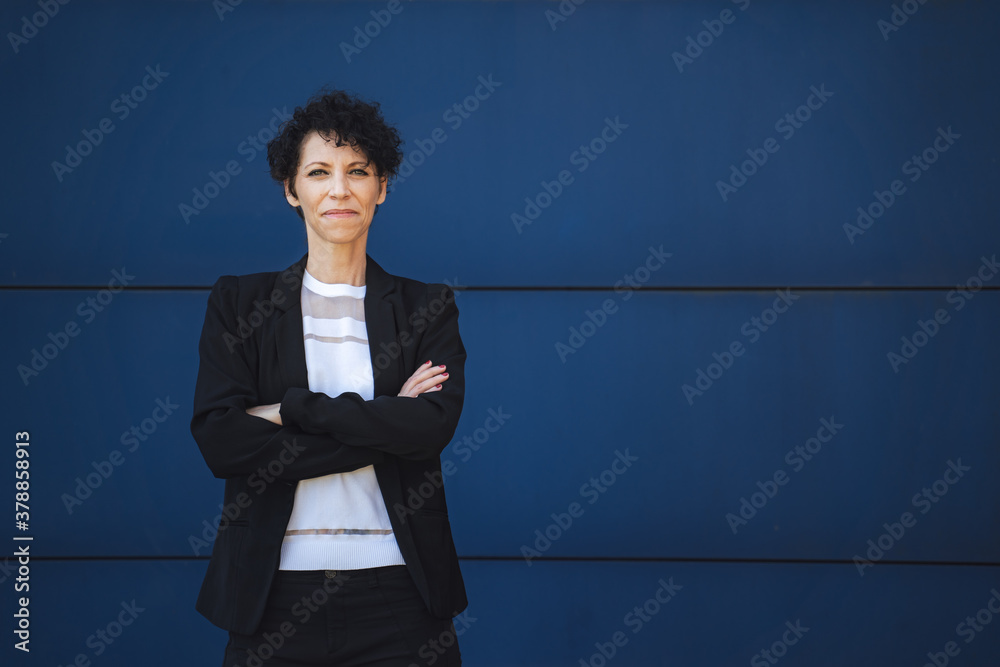 Portrait of confident mature female entrepreneur standing with arms crossed against blue wall - obrazy, fototapety, plakaty 