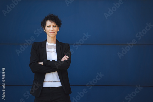 Portrait of confident mature female entrepreneur standing with arms crossed against blue wall