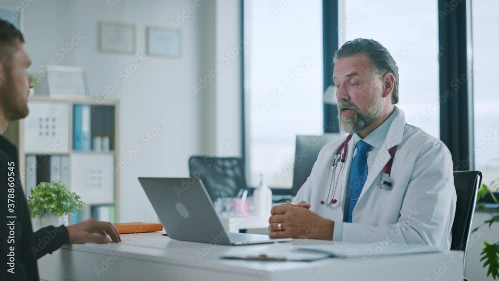 Family Doctor is Delivering Great News About Male Patient's Medical Results During Consultation in a Health Clinic. Physician in White Lab Coat Sitting Behind a Computer in Hospital Office. Family Doctor is Delivering Great News About Male Patient's Medical Results During Consultation in a Health Clinic. Physician in White Lab Coat Sitting Behind a Computer in Hospital Office.