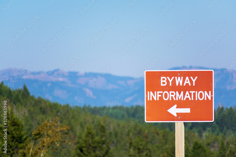Byway information brown sign with directional arrow. Blurred landscape background with forest and mountains