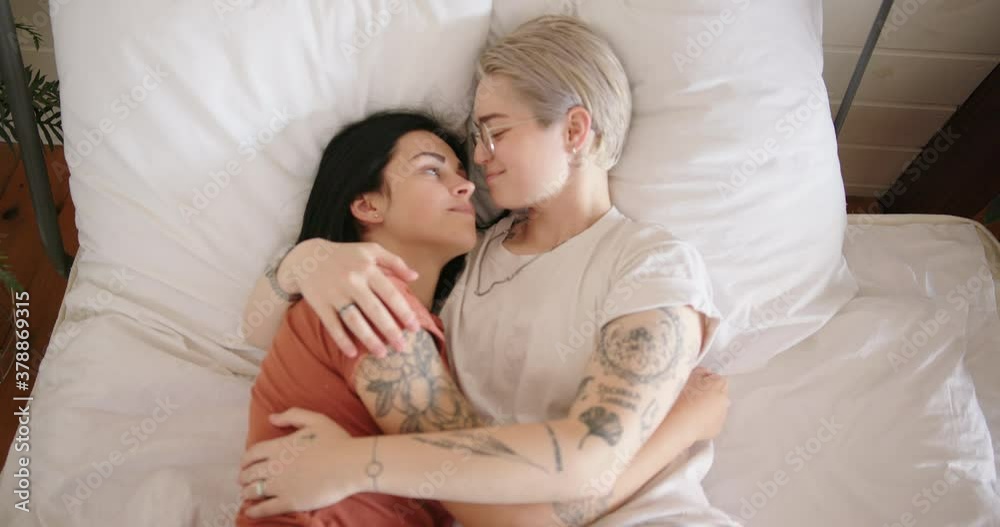 Smiling young woman in white t-shirt and glasses hugs tattooed girlfriend lying on bed between green pot plants slow motion Smiling young woman in white t-shirt and glasses hugs tattooed girlfriend lying on bed between green pot plants slow motion
