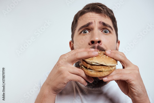A man eating a hamburger on a light background in a white T-shirt cropped view close-up hunger fast food