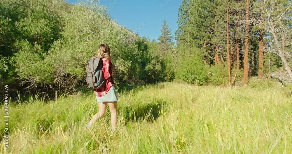 Hiking woman slow motion. Back view of girl in denim skirt, classic red shirt walking with backpack by scenic green grass meadow and forest on background. 4K Outdoor adventure and wild nature travel Hiking woman slow motion. Back view of girl in denim skirt, classic red shirt walking with backpack by scenic green grass meadow and forest on background. 4K Outdoor adventure and wild nature travel