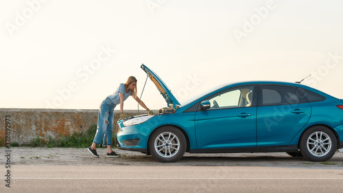 Full length shot of young woman looking under the hood of her broken car, trying to repair it on her own while standing alone after car breakdown on the road side © Svitlana