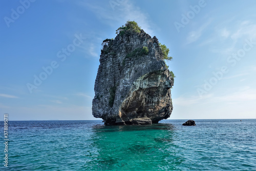 A small uninhabited island in the Andaman Sea against the background of blue sky. A conical rock with steep slopes, few trees at the top. Turquoise water around. Thailand 
